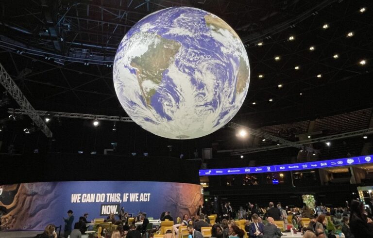 A large globe hangs above a conference hall with attendees seated at tables. A banner reads, "We can do this, if we act now.