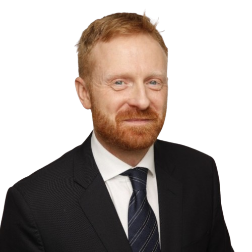 Man with red hair and beard wearing a black suit and striped tie, against a white background.