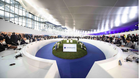 A large, circular conference room with delegates seated around a central grassy area, featuring a screen displaying "G20 Italy 2021.