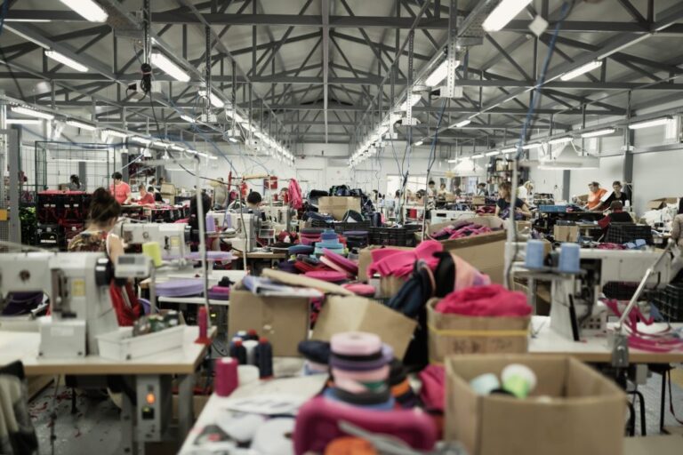 A busy textile factory with sewing machines, fabric rolls, and workers assembling garments. The environment is industrial with overhead lights and scattered materials.