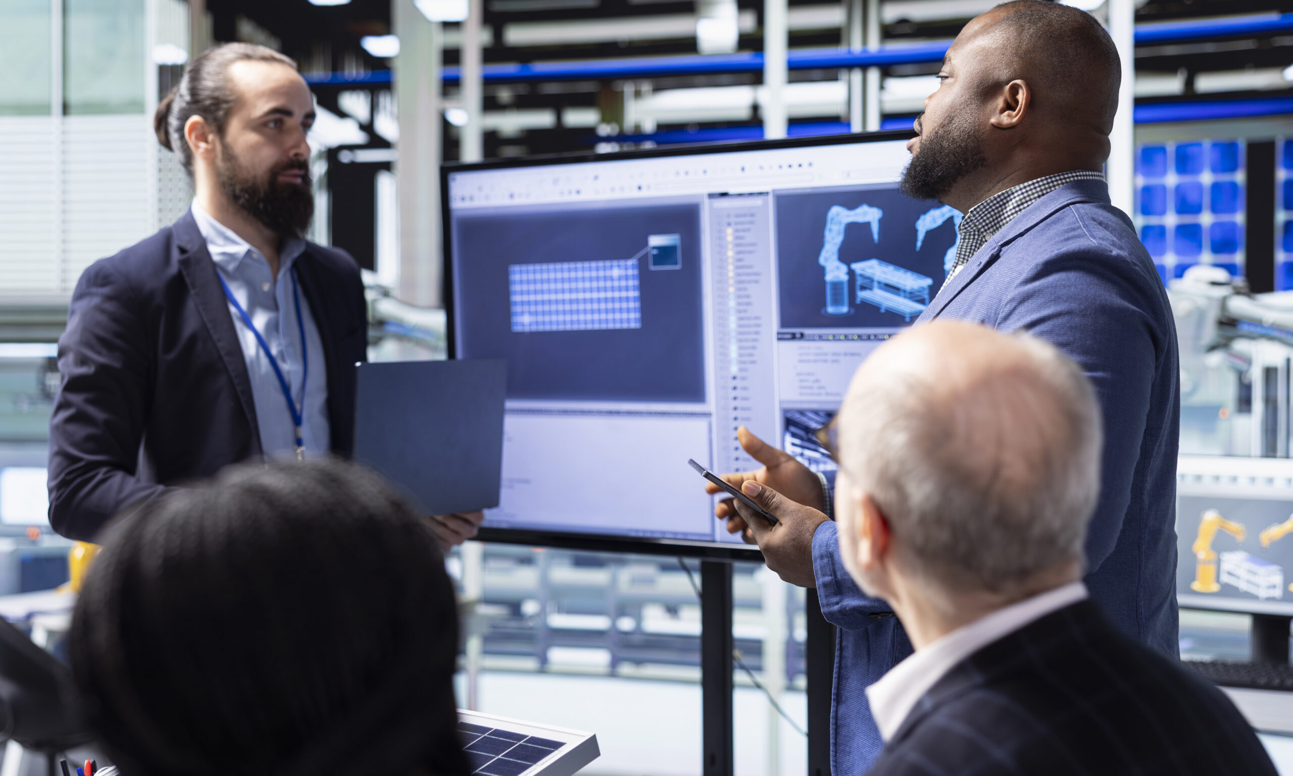 Four people in business attire have a discussion in front of a large screen displaying 3D engineering designs in an office or industrial setting.