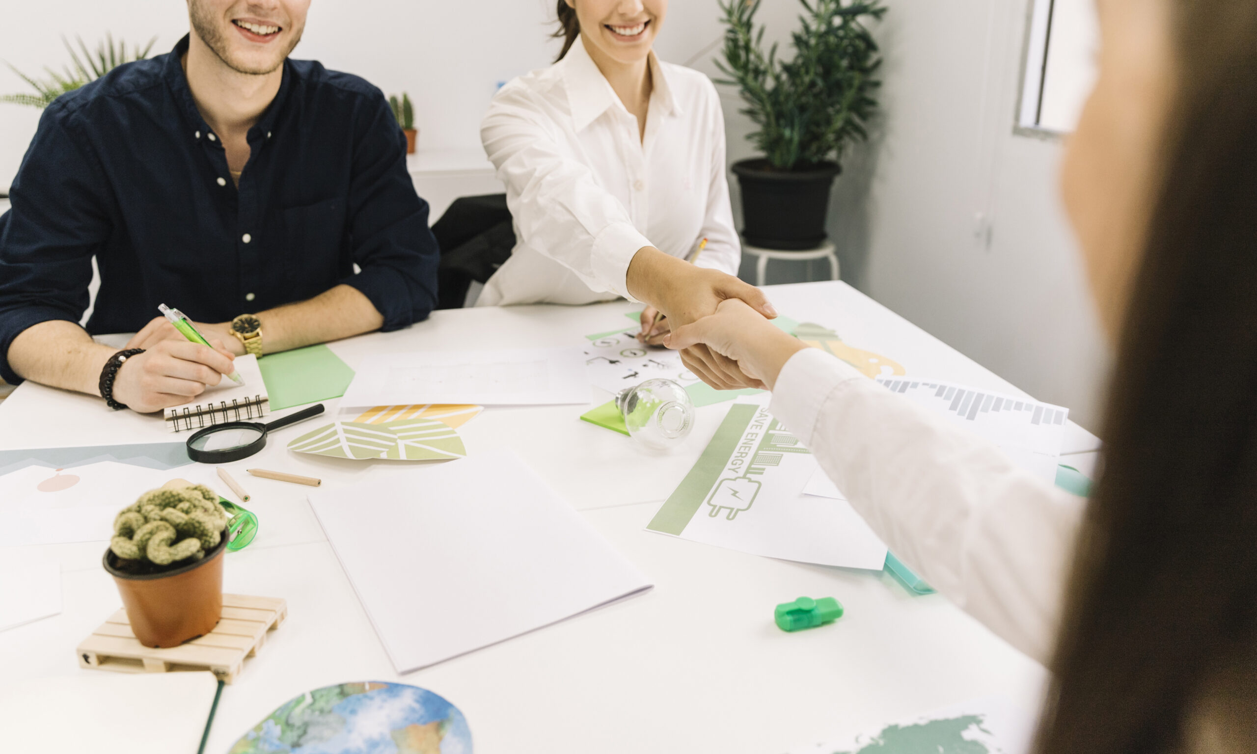 Three people sit at a desk with papers and plants; two of them, a man and a woman, smile as the woman shakes hands with a third person.