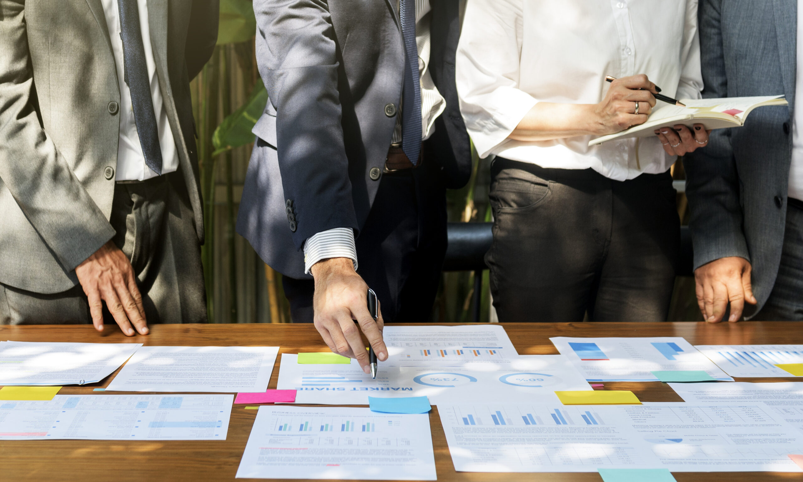 Four people in business attire stand around a table covered with documents, graphs, and charts, discussing and reviewing data. One person points to a paper, another takes notes.