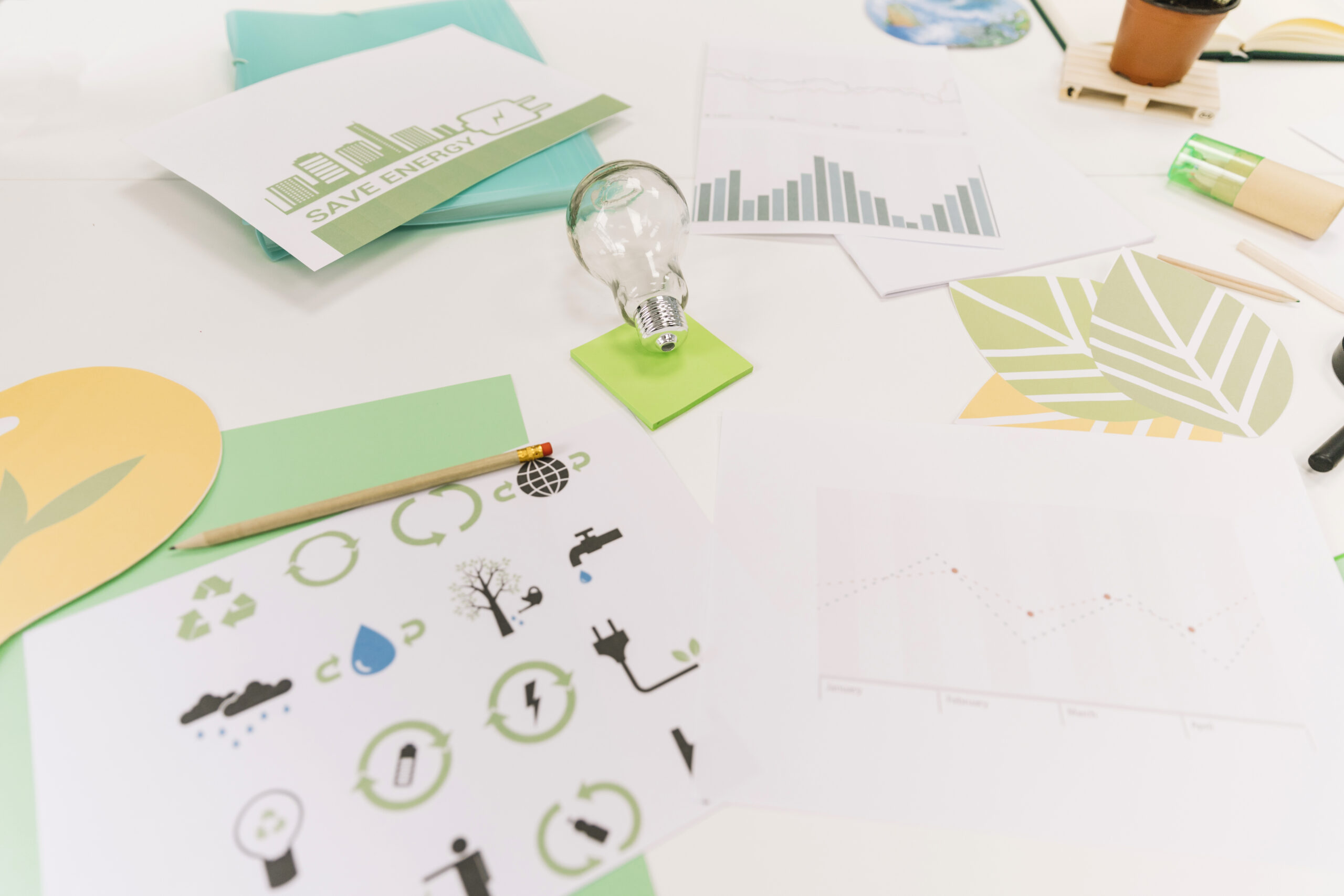 A workspace with charts, graphs, eco-friendly icons, a "Save Energy" flyer, a pencil, and a light bulb on a green stand, emphasizing sustainability and environmental awareness.
