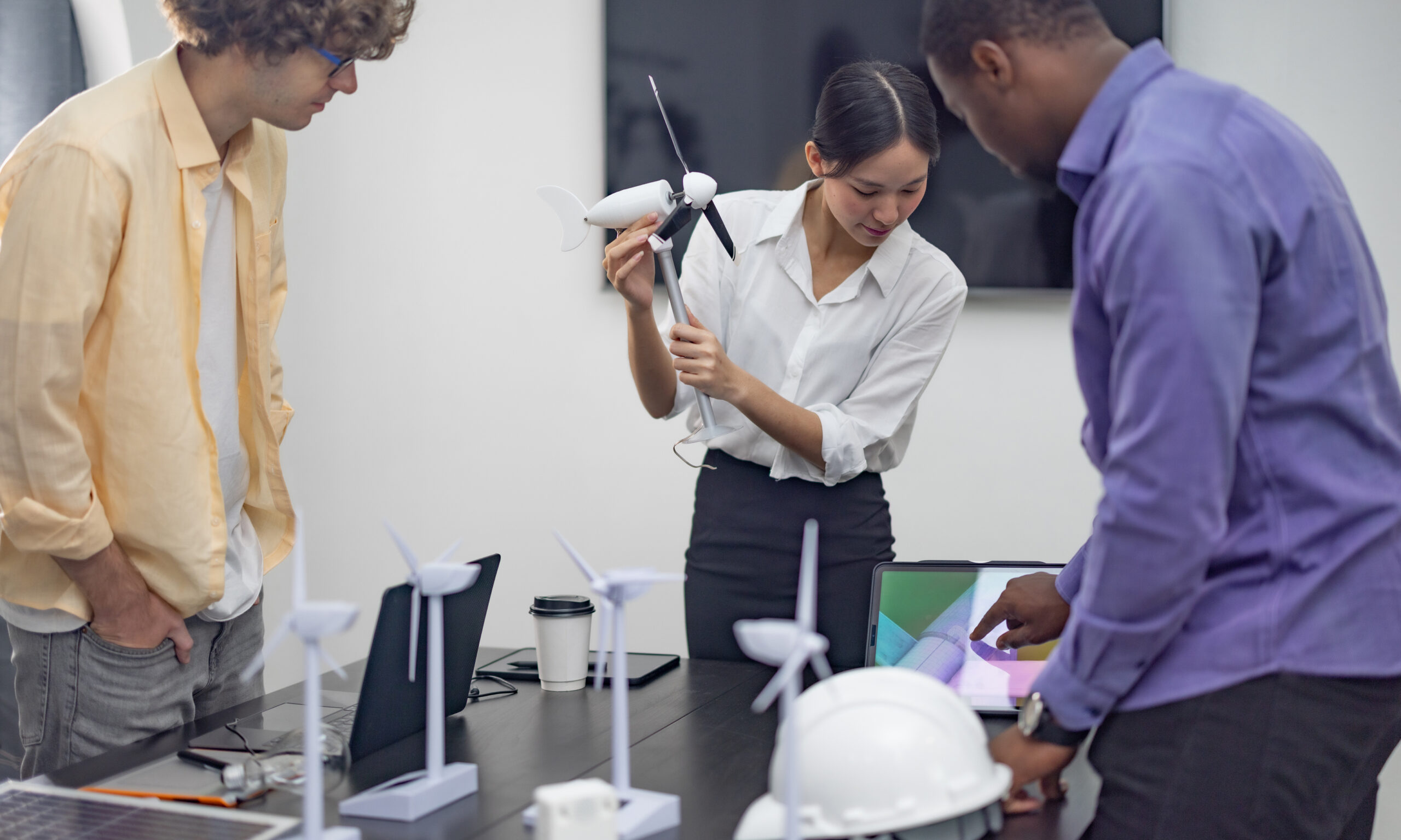 Three people stand around a table with wind turbine models and a laptop, discussing and examining a small wind turbine in a professional setting.